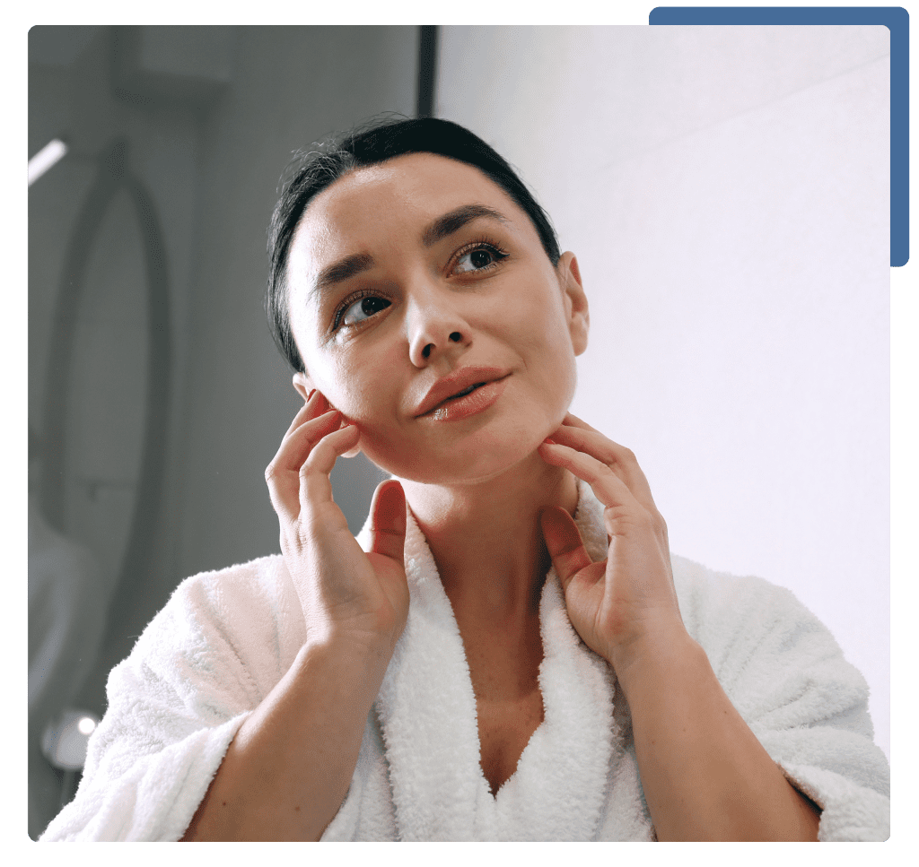 A woman in a bathrobe gazes thoughtfully at her reflection in the mirror, examining her face closely.
